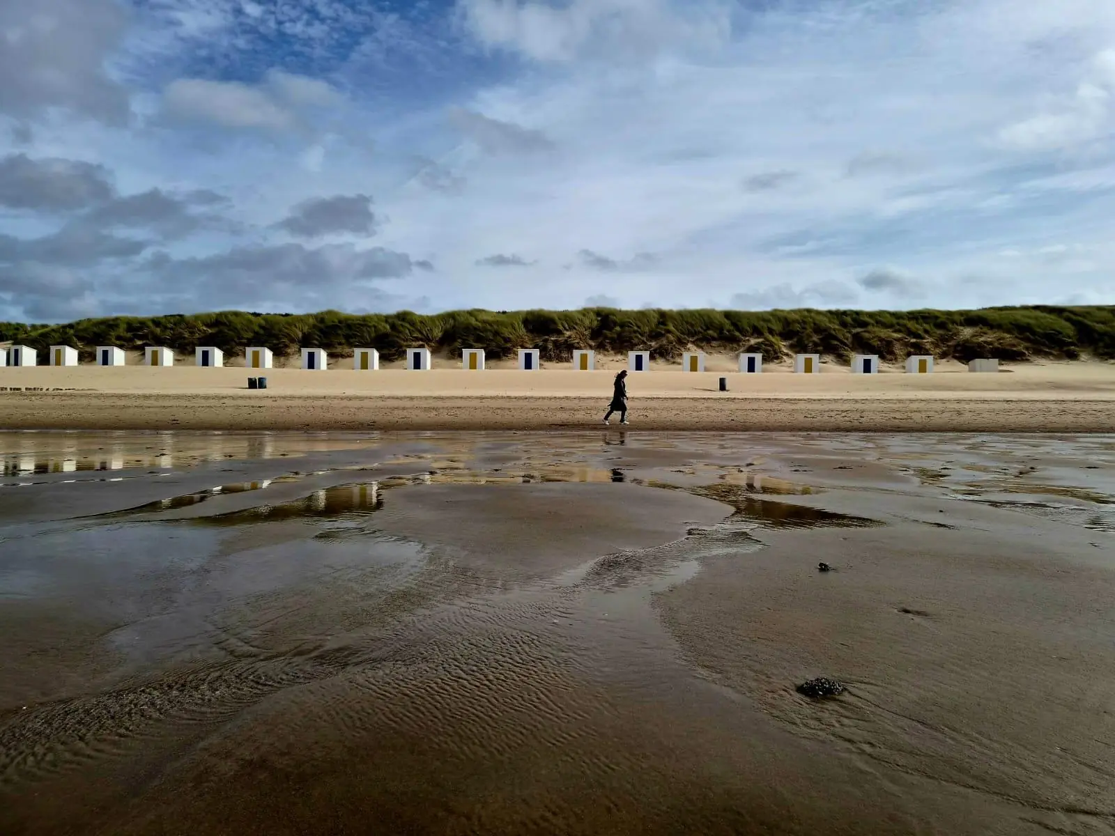 Witte strandhuisjes onder de duinen bij eb, een wandelaar loopt langs